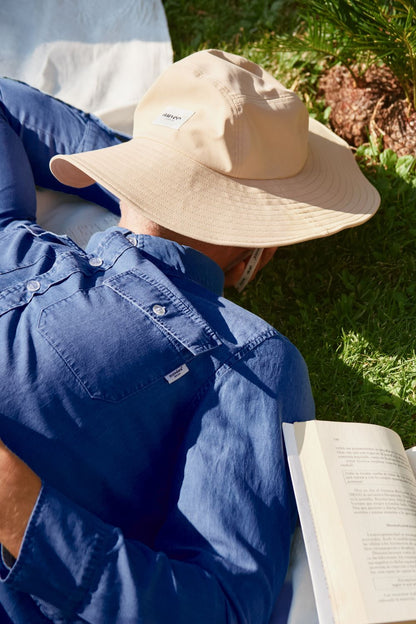 Homme allongé avec un chapeau anti-UV beige, lisant un livre sous le soleil, en tenue décontractée.