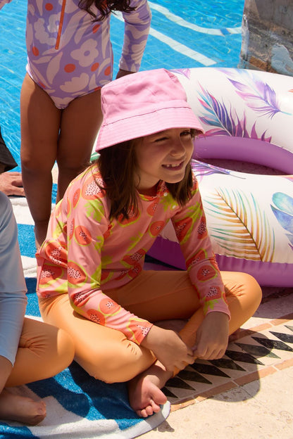 T-shirt de bain anti-UV pour fille, manches longues, UPF 50+, couleur rose et orange, portant un chapeau au bord d'une piscine.